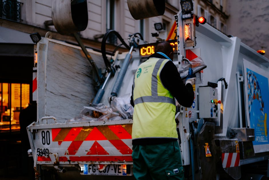 A worker wearing a high-visibility yellow safety vest and dark clothing operates a waste collection vehicle on a city street during dusk. The vehicle has a large, open hopper filled with assorted rubbish, including paper, plastic, and cardboard materials, all appearing loosely piled and ready for disposal. The rear of the truck features red and white reflective chevron markings for visibility, and various mechanical components, hoses, and controls are visible near the operator. The worker's back is turned towards the camera, and they are engaging with the vehicle's controls or monitoring the collection process. In the background, there are illuminated windows of buildings, suggesting an urban environment, with lighting creating a contrast against the darkening sky. The scene reflects an outdoor waste collection operation, exemplifying private or independent rubbish removal services working in a city setting, aligning with services offered by companies like Rubbish Removal Ruislip.