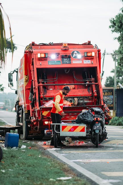 A worker wearing a yellow and red high-visibility vest is operating a red rubbish collection vehicle parked on the side of a street. The vehicle's rear compartment is open, revealing mechanisms and compartments for waste collection. The worker appears to be handling a black plastic bag, possibly preparing to load or unload waste. The scene is set on a paved road with a designated parking or loading area, and there's a small curb with some spilled debris or waste nearby. In the background, there are trees, utility poles, and a building with signage, indicating a semi-urban environment. The environment is well-lit, suggesting daytime, with natural light illuminating the scene. This setup exemplifies private waste collection services, often used as an alternative to council rubbish removal options, with the vehicle and worker actively engaged in rubbish or waste handling.