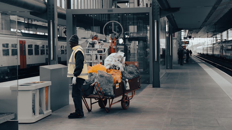 A worker at a modern railway station platform stands beside a large, orange, metal trolley loaded with various waste bags and discarded items, including clear, black, and orange plastic bags filled with rubbish. The worker is dressed in dark clothing, with gloves, and wears a high-visibility vest and a beanie, indicating a professional waste collection role within an infrastructure setting. Behind them, the station features glass panels, metallic structural supports, and a high ceiling with modern lighting, while a train is visible parked on the tracks extending into the distance on the right side of the image. The floor is composed of smooth, grey paving stones, and the overall scene is illuminated by natural and artificial light, creating a neutral, professional atmosphere. The presence of waste bags on the trolley suggests an on-site rubbish collection or disposal process, aligning with private waste handling services such as those offered by Rubbish Removal Ruislip. The environment emphasizes urban waste management functions in a public transportation context, supporting the need for efficient rubbish removal and site clearance in busy transit areas.