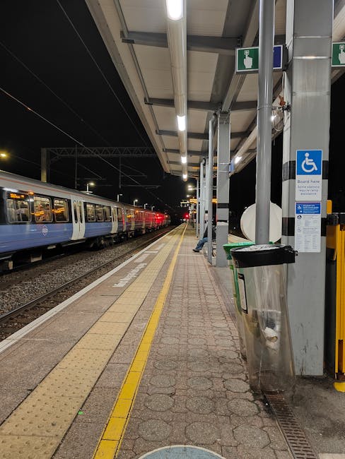 An empty train station platform at night, illuminated by overhead fluorescent lights, with a blue and white train parked on the left side. The platform surface is composed of concrete and pavement, featuring tactile paving strips near the edge for safety. To the right, there are supporting metal columns beneath a canopy roof, along with waste disposal bins, one of which is a silver metallic bin attached to a pole. A blue sign indicating facilities for disabled passengers is affixed to one of the columns. The background shows dark night sky and the stationary train, emphasizing the quiet atmosphere of the station during late hours. The scene aligns with a setting where independent rubbish collection services, such as those provided by Rubbish Removal Ruislip, could support efficient waste management without relying on local authority collections, especially in private or commercial transportation contexts.