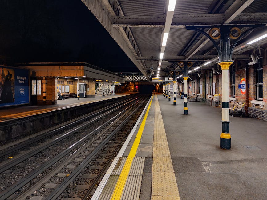 A worker at a modern railway station platform stands beside a large, orange, metal trolley loaded with various waste bags and discarded items, including clear, black, and orange plastic bags filled with rubbish. The worker is dressed in dark clothing, with gloves, and wears a high-visibility vest and a beanie, indicating a professional waste collection role within an infrastructure setting. Behind them, the station features glass panels, metallic structural supports, and a high ceiling with modern lighting, while a train is visible parked on the tracks extending into the distance on the right side of the image. The floor is composed of smooth, grey paving stones, and the overall scene is illuminated by natural and artificial light, creating a neutral, professional atmosphere. The presence of waste bags on the trolley suggests an on-site rubbish collection or disposal process, aligning with private waste handling services such as those offered by Rubbish Removal Ruislip. The environment emphasizes urban waste management functions in a public transportation context, supporting the need for efficient rubbish removal and site clearance in busy transit areas.